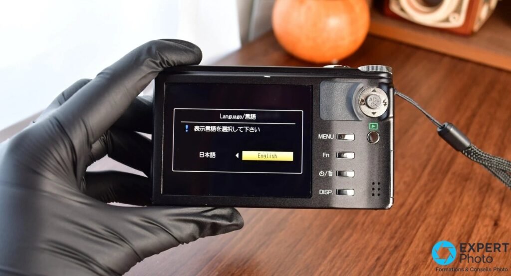  Dos du Ricoh CX3 tenu en main gantée noire, écran affichant le menu de sélection de langue Japanese/English 