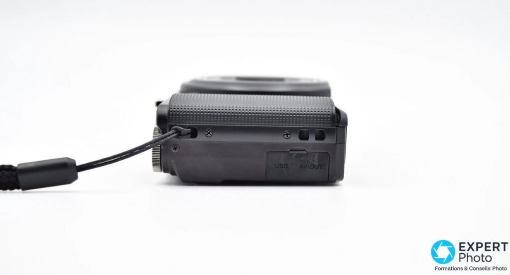 Ricoh CX3 vu de profil avec trappe USB et AV-OUT visible, dragonne noire attachée à l'anneau gauche 