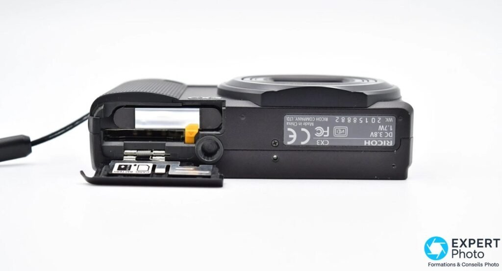 Ricoh CX3 vu de dessous avec trappe batterie ouverte et étiquette CE/FCC