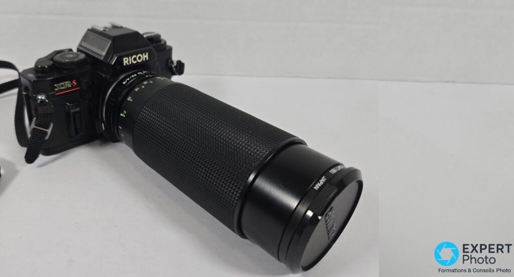 Reflex Ricoh XR-S noir avec long téléobjectif zoom monté en monture K, vue 3/4 avant et bouchon d'objectif clipsé.