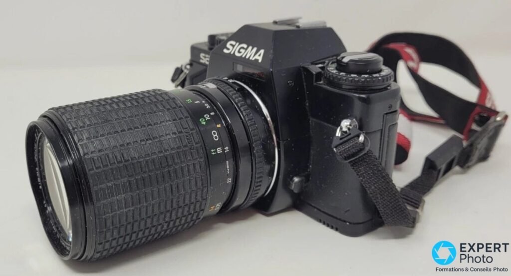 Sigma SA-1 vu en trois-quarts côté objectif, zoom Sigma 35-135 mm déployé en position télé, sangle apparente.