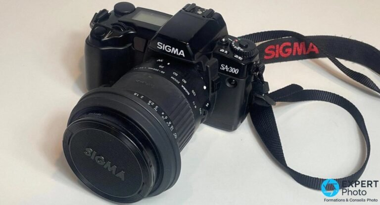 Sigma SA-300 noir vu de trois quarts avant, zoom Sigma 28-200 mm avec bouchon SIGMA et sangle SIGMA rouge