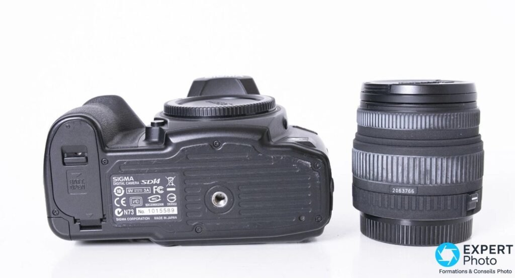 Sigma SD14 vu de dessous avec plaque constructeur Sigma SD14 et numéro de série lisible, objectif Sigma posé verticalement à droite.