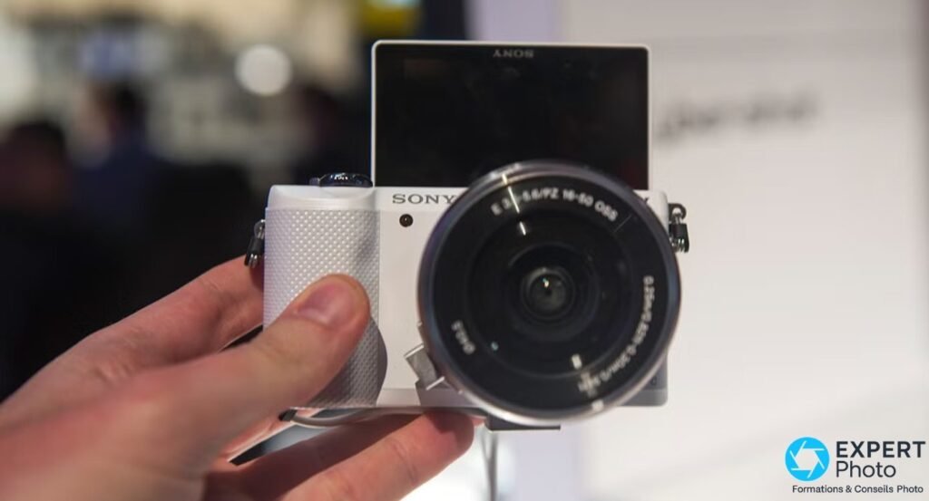 Sony A5000 blanc tenu en main, écran relevé à 180° vers le haut, position autoportrait, zoom 16-50 monté.