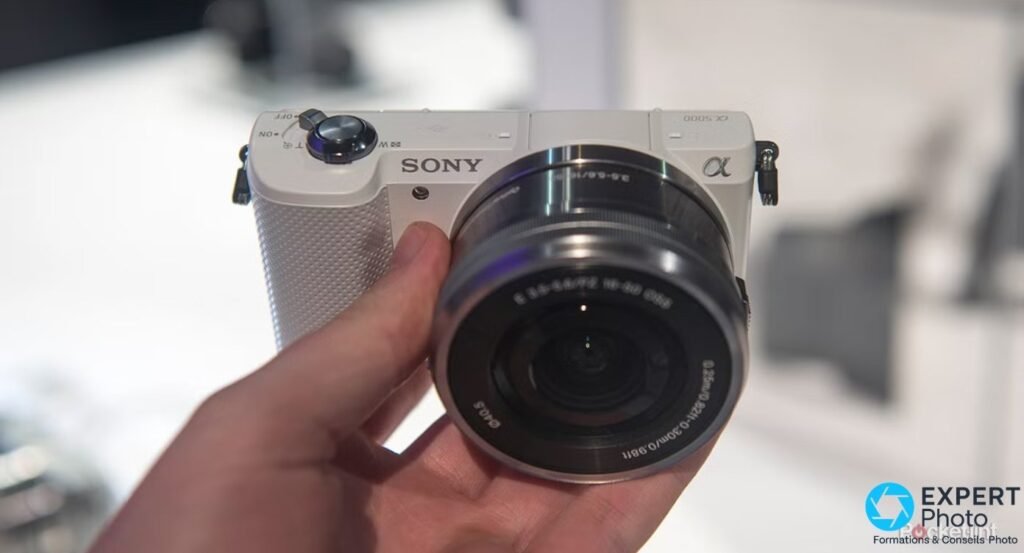 Sony A5000 blanc tenu en main, vue de face, logo SONY et α 5000 visibles, zoom 16-50 PZ OSS monté.