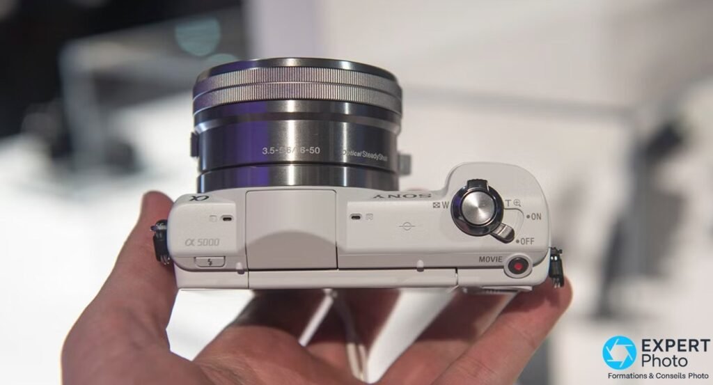 Sony A5000 blanc tenu en main, vue de dessus, zoom 16-50 monté, bouton MOVIE et levier ON/OFF visibles. 