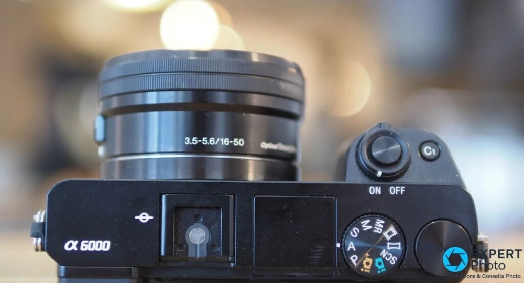 Sony A6000 vu de dessus, griffe flash, molette M/MR/S/A/P/SCN, objectif 16-50 mm vu de côté.