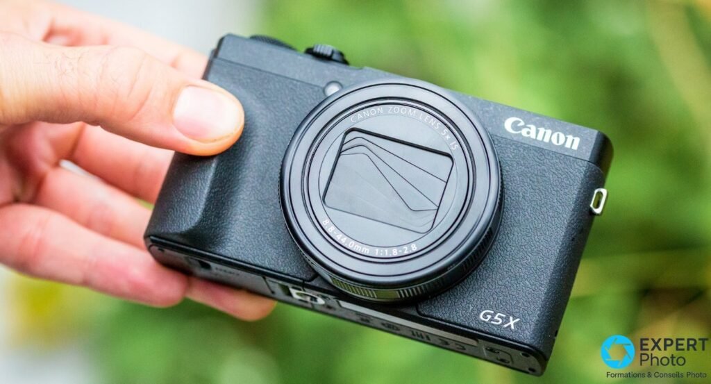 Canon G5 X Mark II tenu en main avec objectif rétracté et cache d'objectif fermé, vue frontale sur bokeh vert.