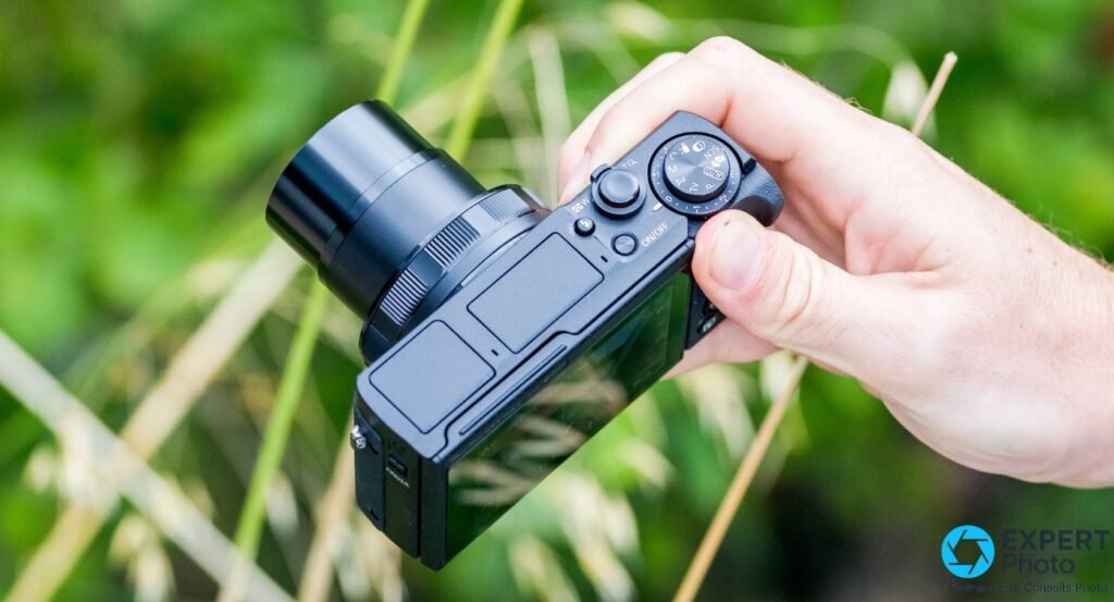 Canon G5 X Mark II tenu en main, vue 3/4 dessus avec objectif déployé et molette des modes, fond herbes vertes. 
