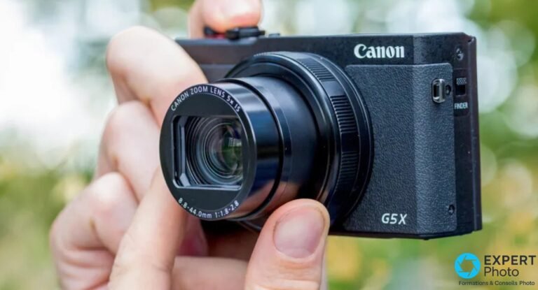Canon G5 X Mark II tenu en main avec objectif Canon Zoom Lens 5x IS déployé, vue 3/4 avant sur fond végétation floue.