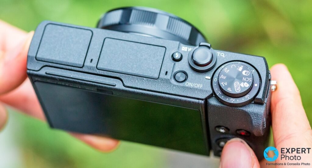 Canon G5 X Mark II vue du dessus, molette des modes M Av Tv P AUTO SCN et bouton ON/OFF visibles, tenu en main. 