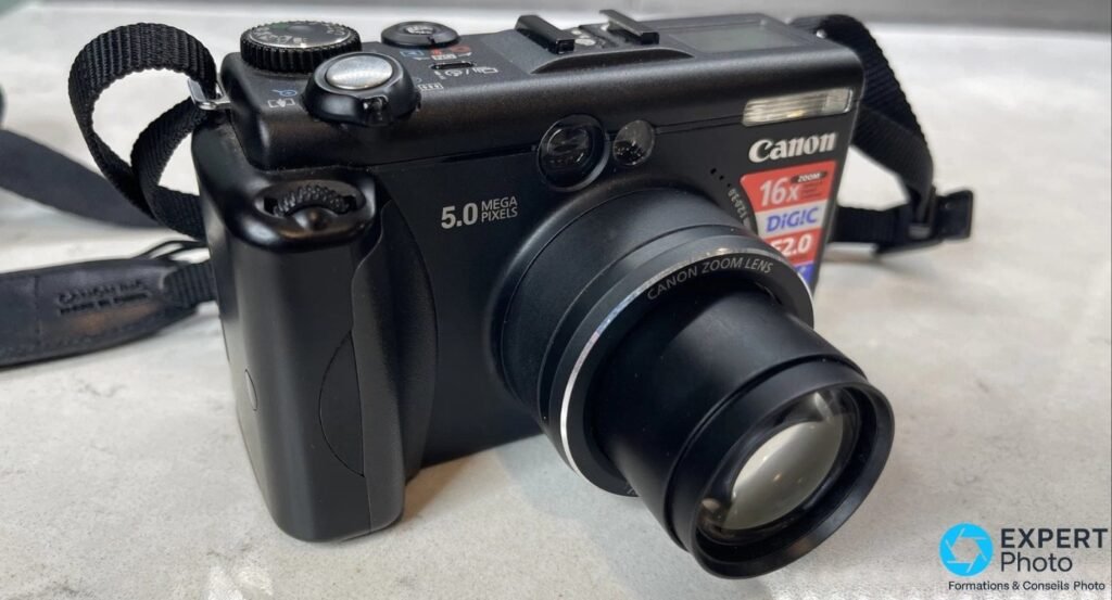 Canon PowerShot G5 noir en trois-quarts avant gauche, objectif Canon Zoom Lens déployé en téléobjectif, grip visible.