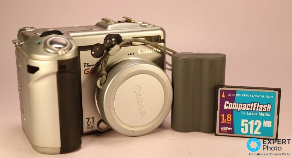 Canon PowerShot G6 avec bouchon d'objectif Canon, batterie BP-511A et carte CompactFlash Lexar 512 Mo sur fond rose pâle.