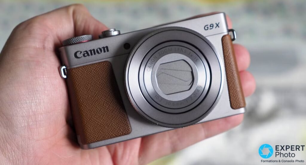 Canon PowerShot G9 X Mark II argenté tenu en main, vue avant frontale, volet objectif fermé, logos Canon et G9 X visibles.