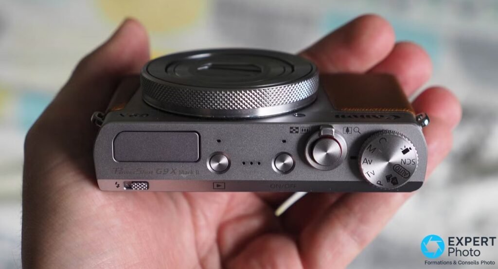 Canon PowerShot G9 X Mark II tenu en main, vue de dessus avec gravure du modèle et molette de mode visibles. 