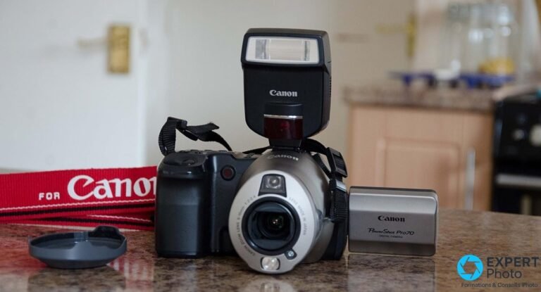 Canon PowerShot Pro70 en 2026 : avis, achat d’occasion et alternatives Canon PowerShot Pro70 vu de trois-quarts avant, flash externe Canon, sangle, cache et boîte d'origine sur plan en granit.