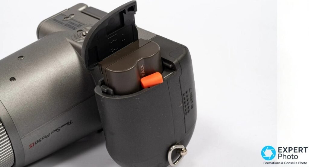 Canon PowerShot Pro90 IS, compartiment batterie ouvert sur le grip, batterie partiellement insérée, languette orange.