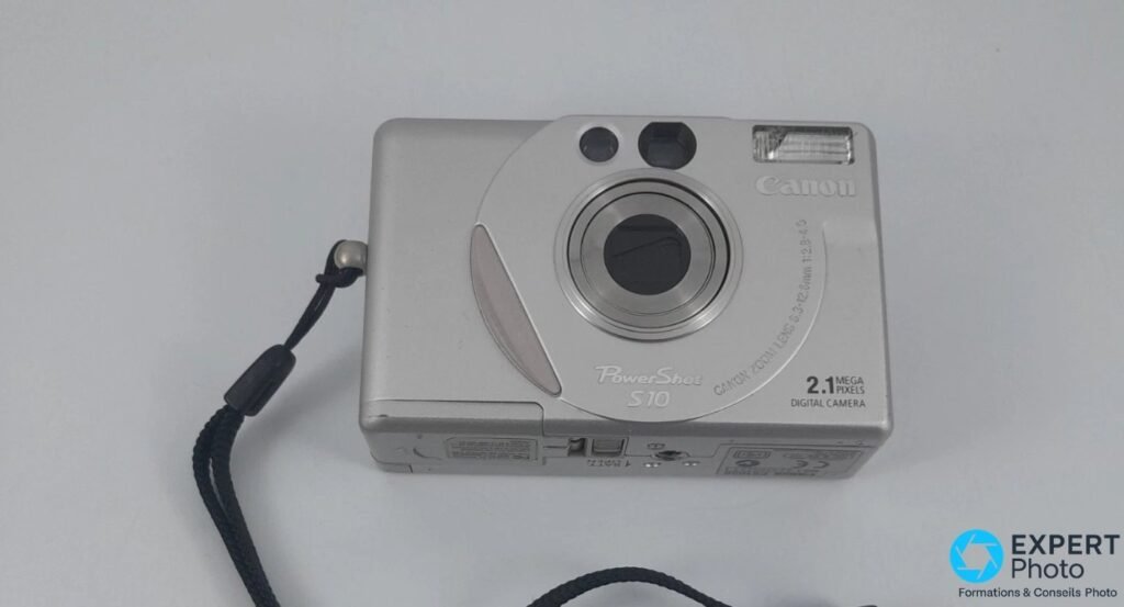 Canon PowerShot S10 vu de trois-quarts face avec sa dragonne noire déroulée à gauche du boîtier.