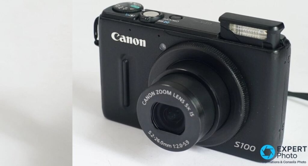 Canon PowerShot S100 vu en trois quarts avant, flash pop-up sorti et objectif sorti, logo Canon et S100 visibles.