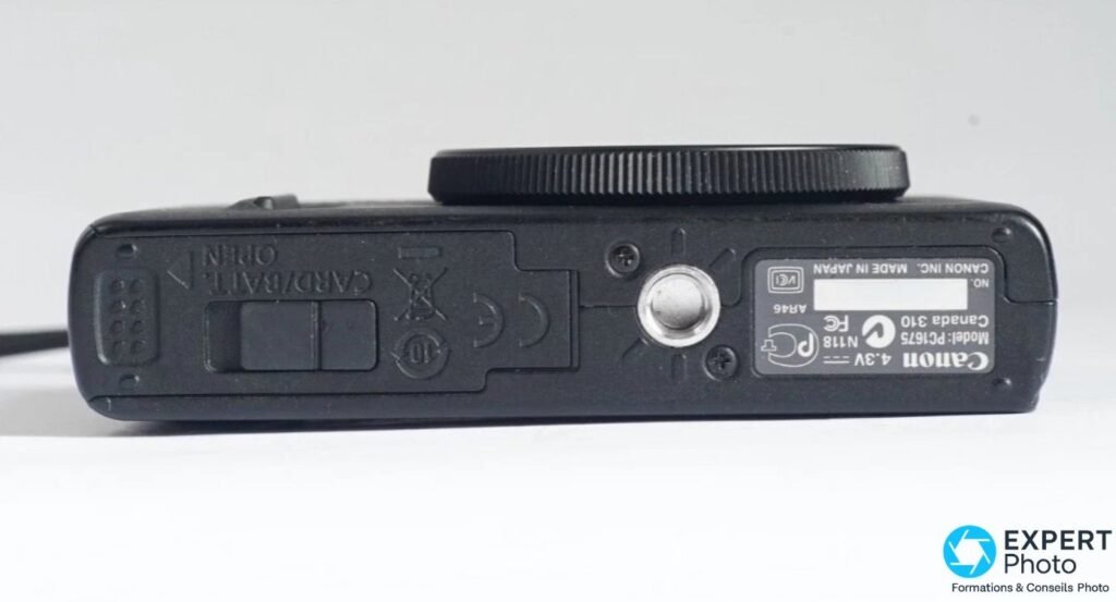 Canon PowerShot S100 vu de dessous, étiquette Canon Model PC1675 Made in Japan et trappe carte/batterie.