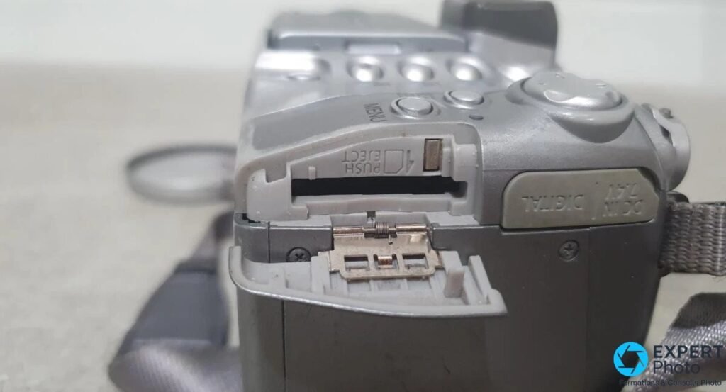 Canon PowerShot S2 IS avec compartiment piles AA et logement carte SD ouverts, marquage Push Eject visible et port DC IN 7.4V.