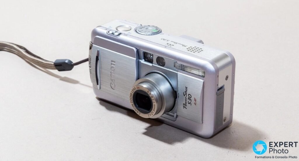 Canon PowerShot S30 silver vu de trois quarts avant sur fond clair, objectif zoom 7,1-21,3 mm sorti, dragonne grise visible. 