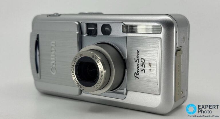 Test Canon PowerShot S50 : faut-il l’acheter d’occasion en 2026 ? Canon PowerShot S50 argenté de face en 3/4 droite, capot ouvert, objectif Canon Zoom Lens 3x déployé, viseur visible.