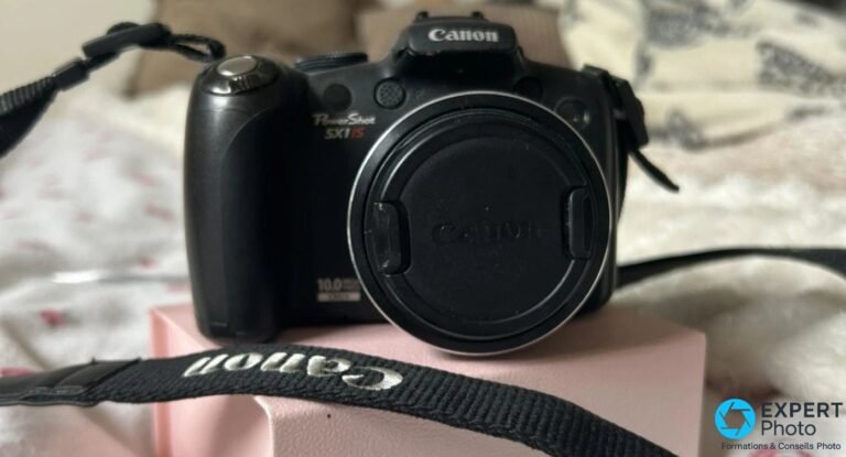 Canon PowerShot SX1 IS : test terrain et avis en 2026 Canon PowerShot SX1 IS vu de face, marquages PowerShot SX1 IS et 10 mégapixels CMOS, bouchon en place, bandoulière Canon.