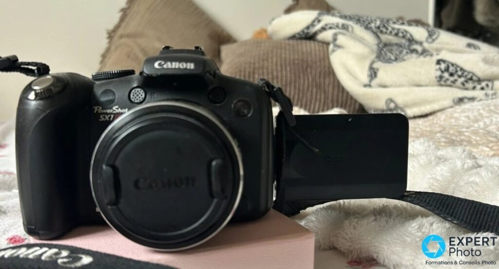 Canon PowerShot SX1 IS vu de face, bouchon en place, écran orientable LCD ouvert et basculé à plat sur le côté droit.