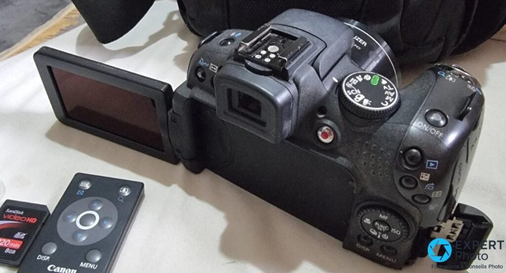 Canon PowerShot SX1 IS de trois quarts arrière, écran déployé, télécommande sans fil et carte SD SanDisk Video HD à côté.
