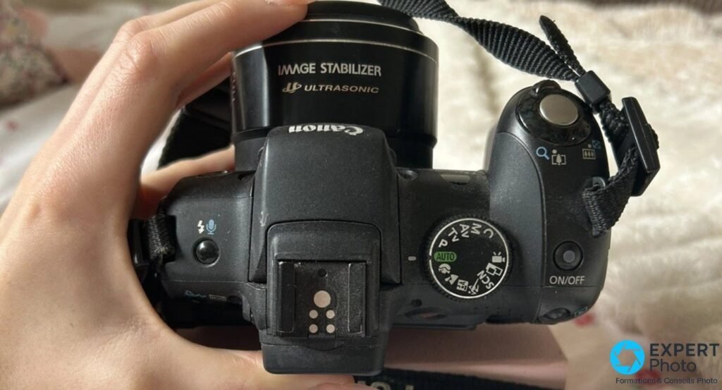 Canon PowerShot SX1 IS vu de dessus, objectif Image Stabilizer Ultrasonic, molette PASM, griffe flash et bouton ON/OFF.
