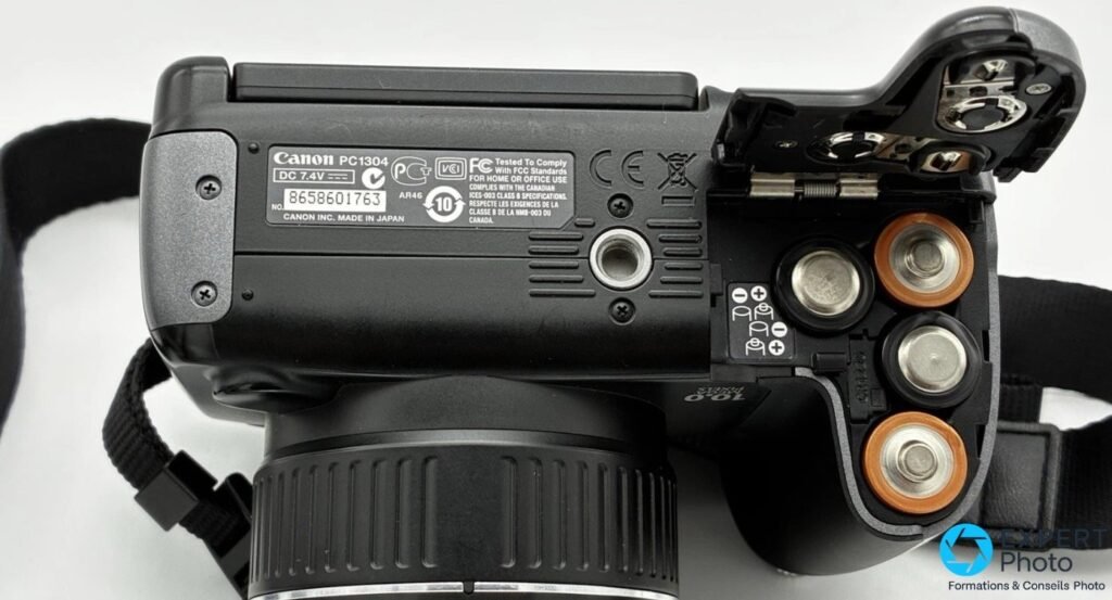 Canon PowerShot SX10 IS vue de dessous, compartiment piles ouvert avec 4 piles AA, plaque « Canon PC1304 DC 7.4V »