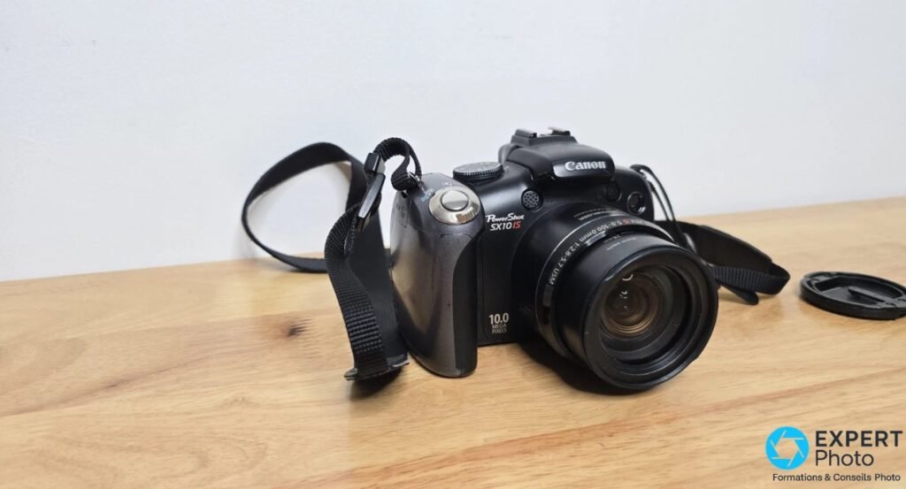 Canon PowerShot SX10 IS sur table bois, vue 3/4 avant, objectif Canon zoom 20× IS 5.0-100mm sorti, bouchon à droite