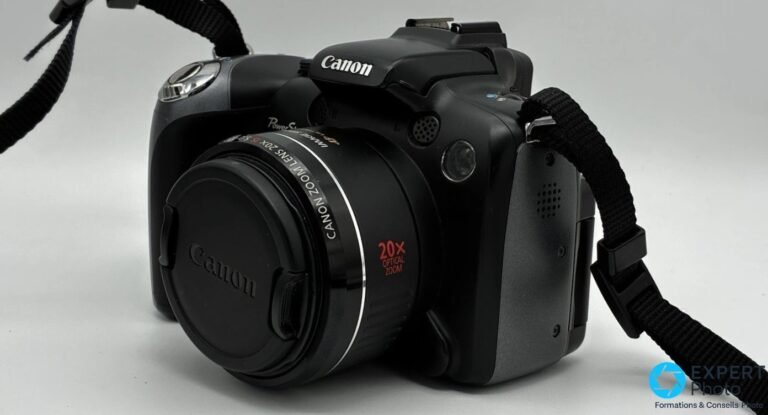 Canon PowerShot SX10 IS : test terrain du bridge 20× Canon PowerShot SX10 IS noir, vue 3/4 avant, bouchon objectif Canon en place, anneau « 20x optical zoom », fond studio gris