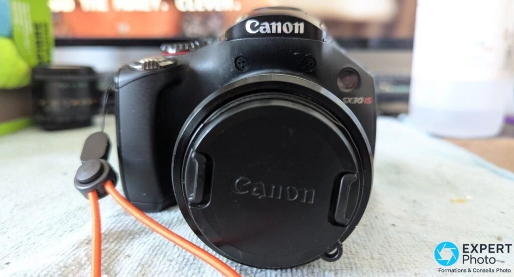 Face frontale du Canon PowerShot SX30 IS, optique rétractée avec capuchon Canon, logo SX30 IS rouge visible sur la lunette.