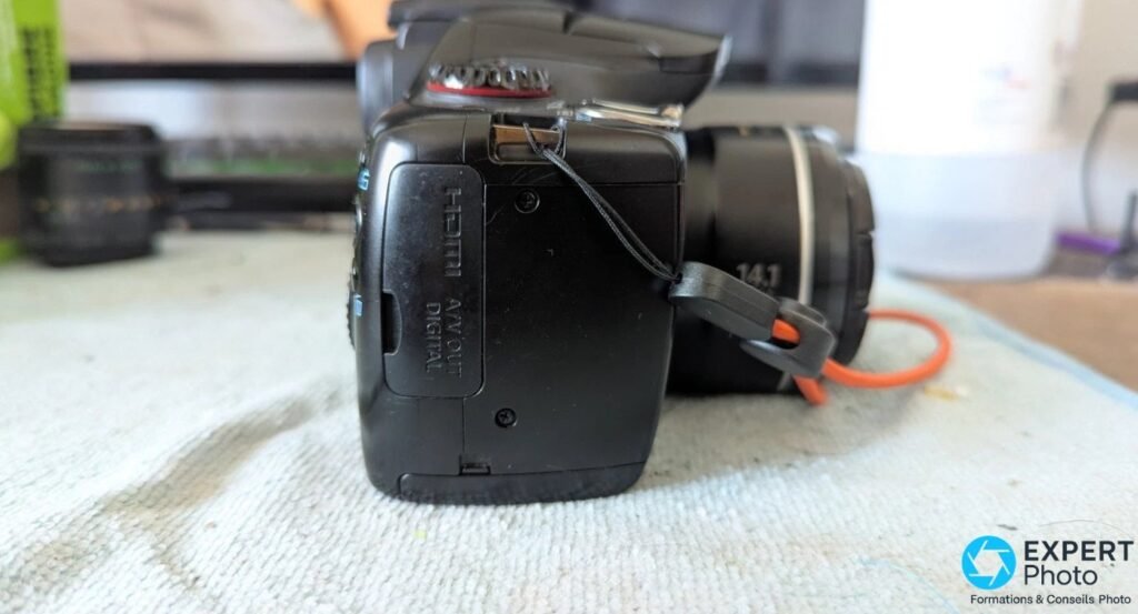 Profil droit du Canon PowerShot SX30 IS posé sur tapis clair, trappe de ports HDMI fermée et dragonne orange suspendue.