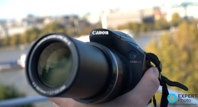 Canon PowerShot SX40 HS tenu à une main, zoom 35x déployé, devant un paysage urbain flou avec arbres d'automne.