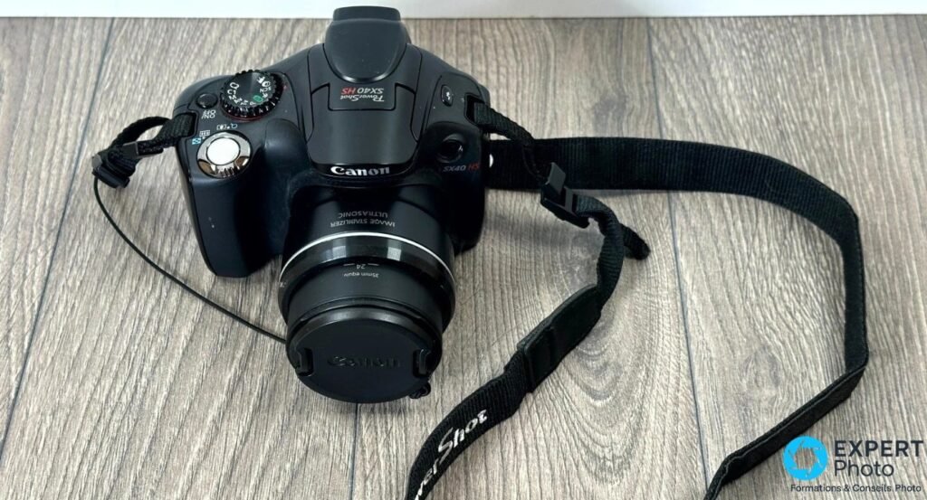 Canon PowerShot SX40 HS noir posé à plat sur fond bois, bouchon Canon en place et dragonne PowerShot étalée. 
