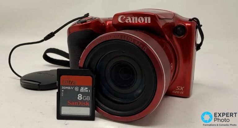 Canon PowerShot SX420 IS rouge, vue 3/4 avant, objectif Canon Zoom Lens 42x IS sorti, cache et carte SD SanDisk 8 Go au sol