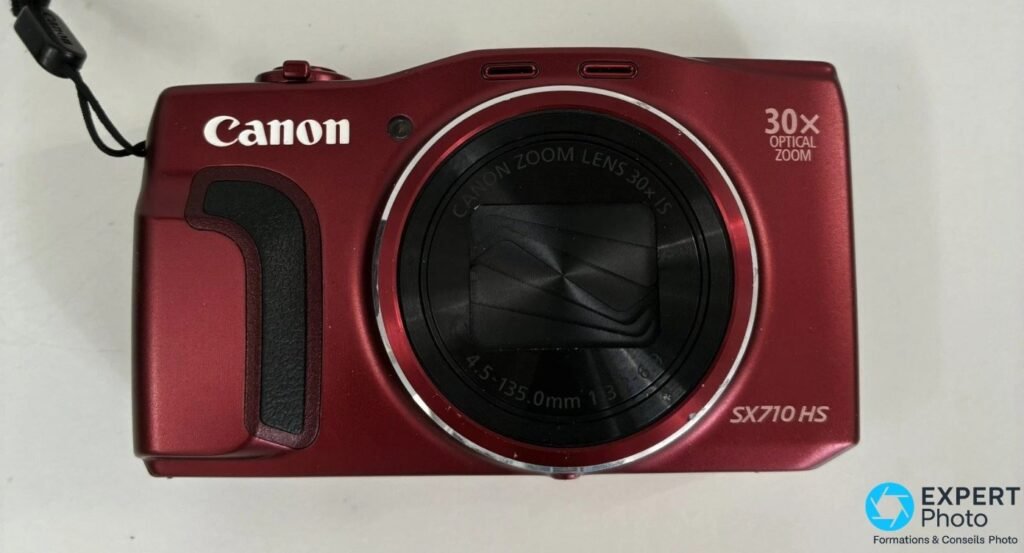 Canon PowerShot SX710 HS rouge de face, objectif rétracté avec volet fermé et mention 30x Optical Zoom gravée. 