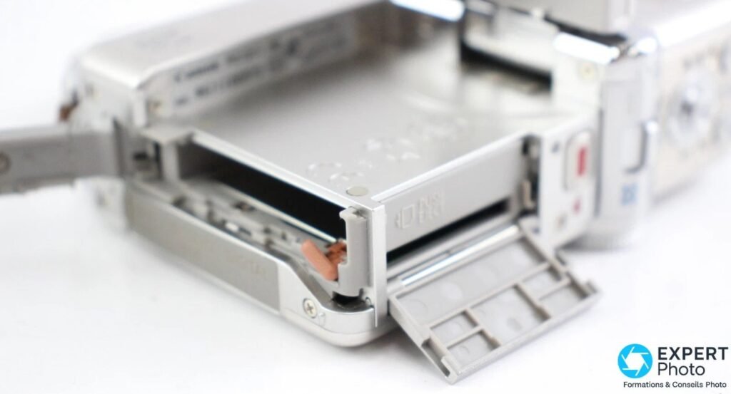 Canon PowerShot TX1 posé à plat avec trappe carte SD et batterie ouverte, ressort de verrouillage visible