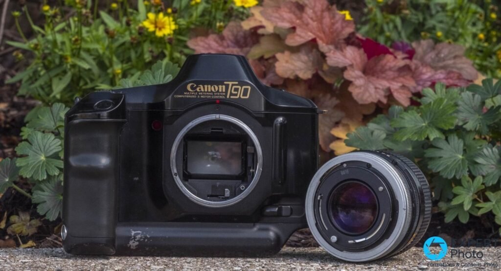 Canon T90 boîtier nu avec monture FD apparente, objectif Canon FD posé debout à côté, fond fleuri en arrière-plan