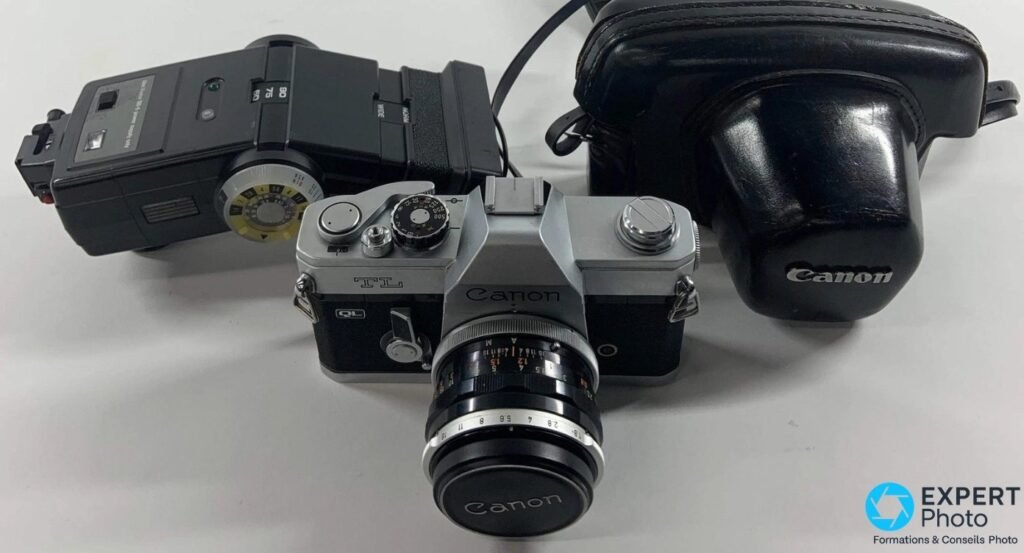 Canon TL QL 1968 vue 3/4 face, flash cobra noir à gauche, étui cuir noir gravé Canon à droite, fond blanc.