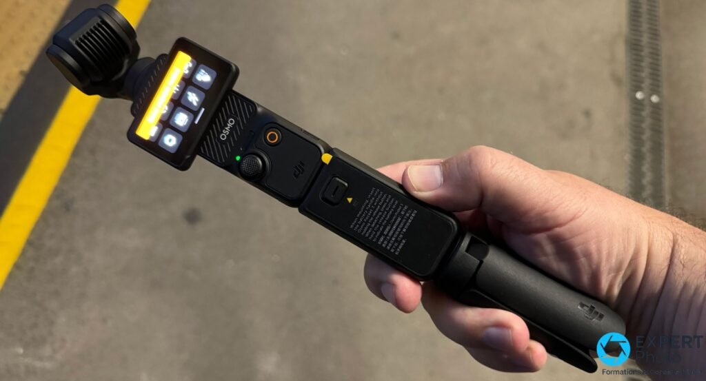 DJI Osmo Pocket 3 avec poignée batterie DJI en main, marquage OSMO visible, écran orange allumé, usage extérieur