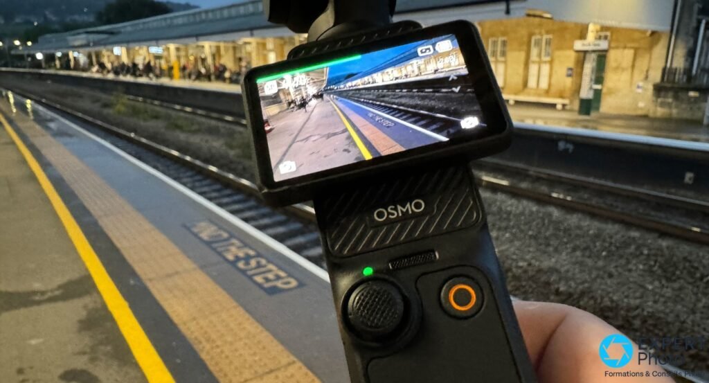 DJI Osmo Pocket 3 tenue en main, écran retourné en mode selfie, aperçu d'un quai de gare de nuit sur l'écran OLED