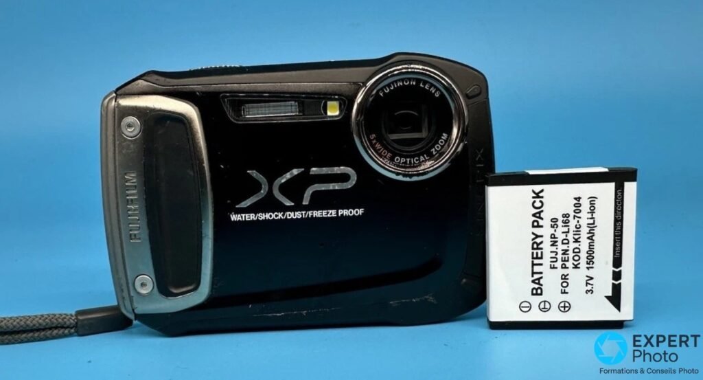 Fujifilm FinePix XP100 noir vu de face, objectif Fujinon zoom 5x rétracté, à côté d'une batterie compatible NP-50.