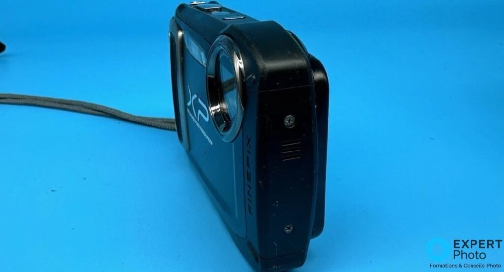 Fujifilm FinePix XP100 vu de trois quarts arrière droit, gravure FINEPIX et grille latérale visibles.