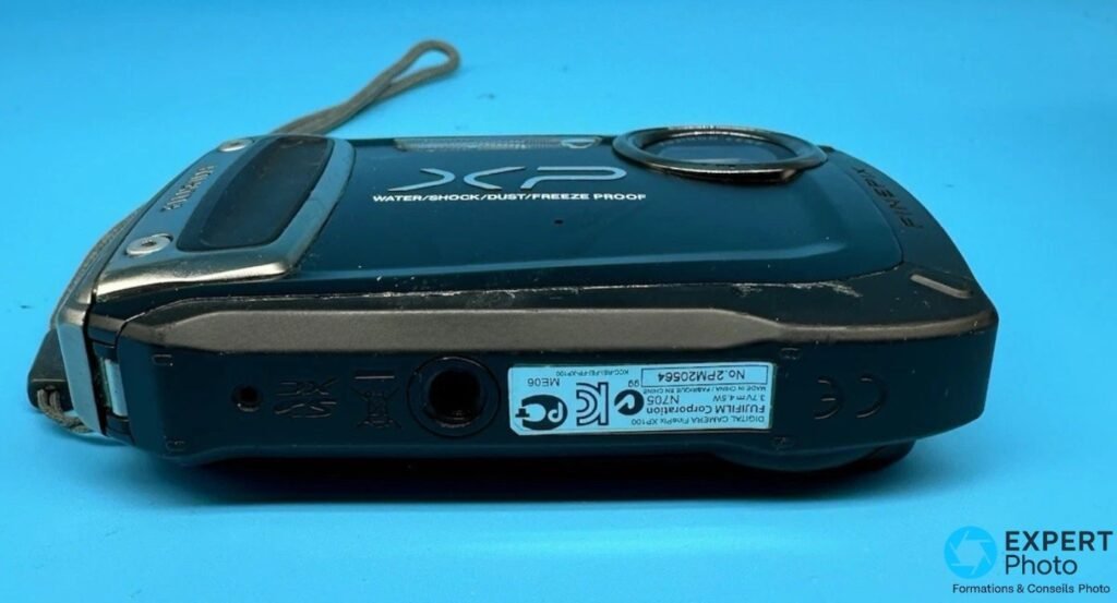 Fujifilm FinePix XP100 vu de dessous, étiquette FUJIFILM Corporation, embase trépied et trappe batterie SD.
