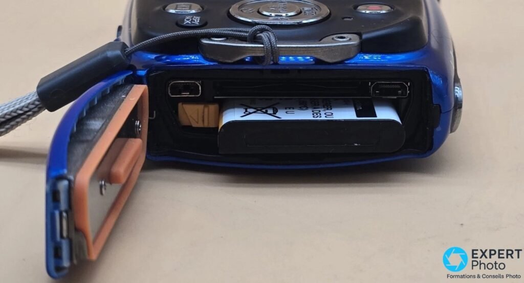 Compartiment batterie du Fujifilm FinePix XP50, trappe latérale ouverte avec joint d'étanchéité orange visible.