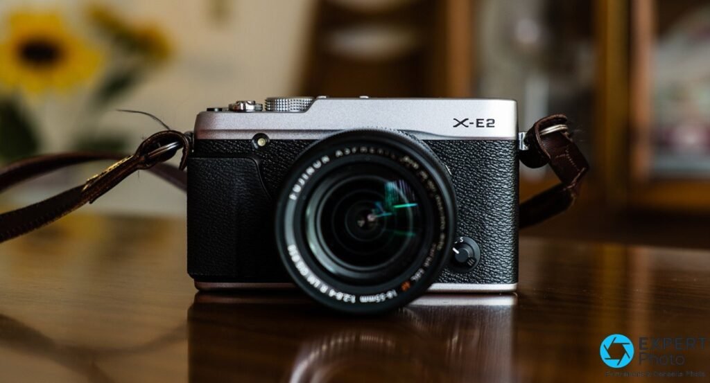  Fujifilm X-E2 silver de face avec Fujinon XF 18-55mm f/2.8-4 R LM OIS, sangle cuir bordeaux étalée, sur table en bois sombre. 
